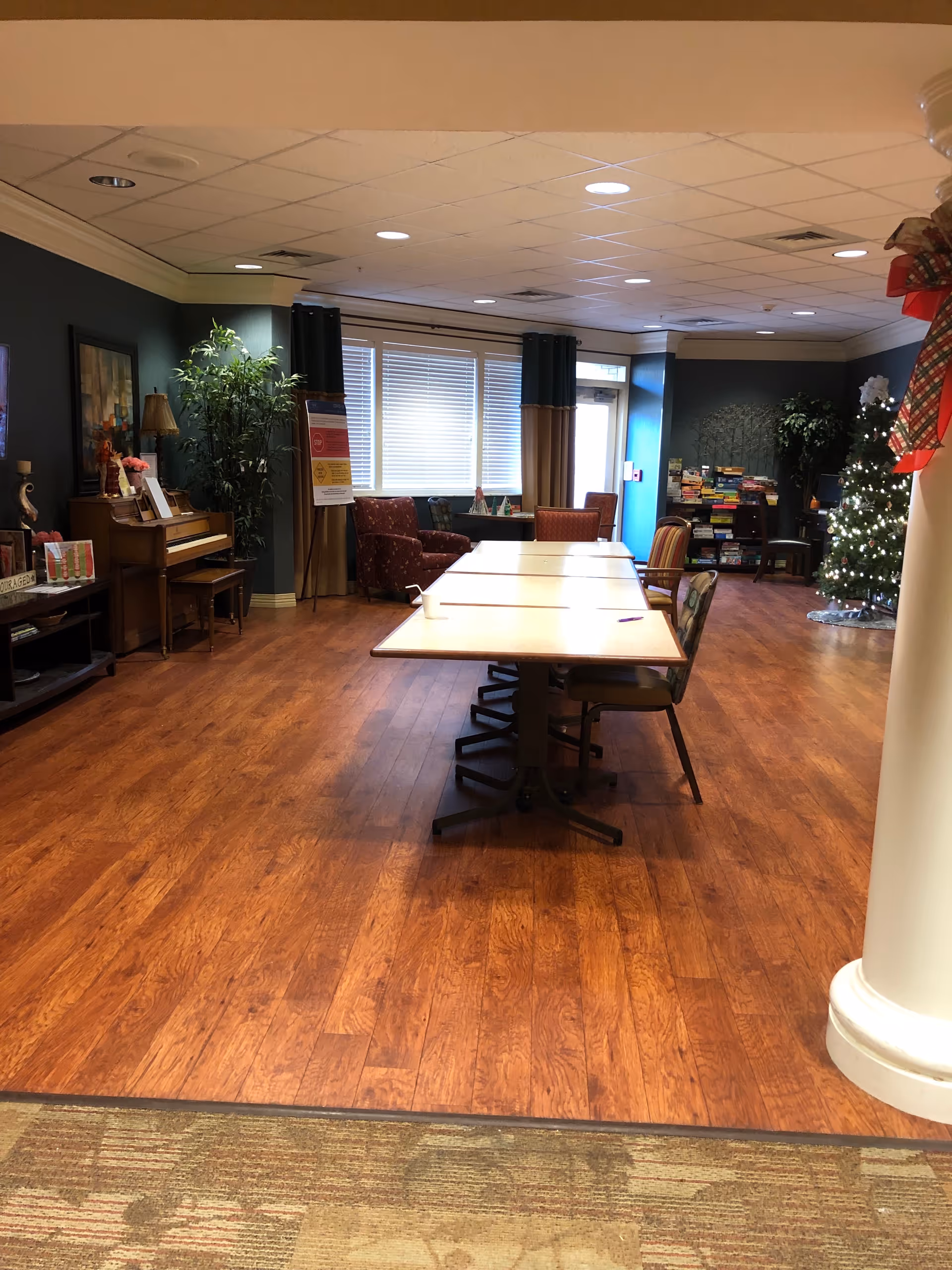 Interior common room with long tables, chairs, a piano, plants, and a Christmas tree.