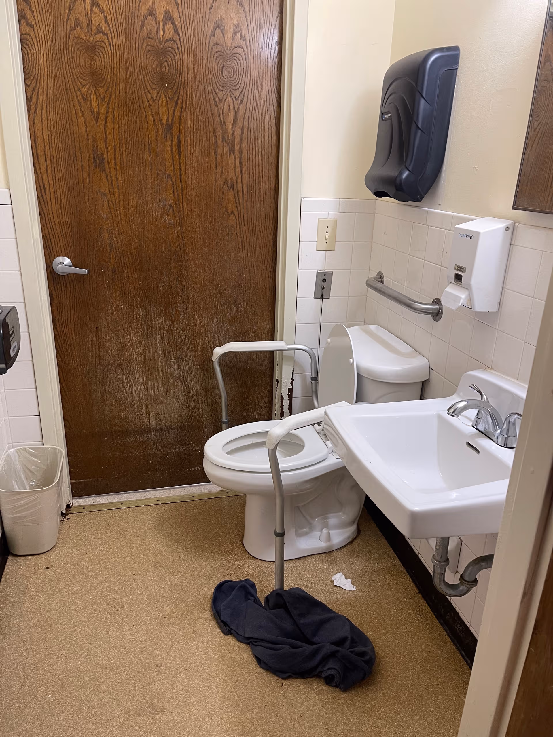 Accessible bathroom featuring toilet, sink, grab bars, soap dispenser, and a dark cloth on floor.