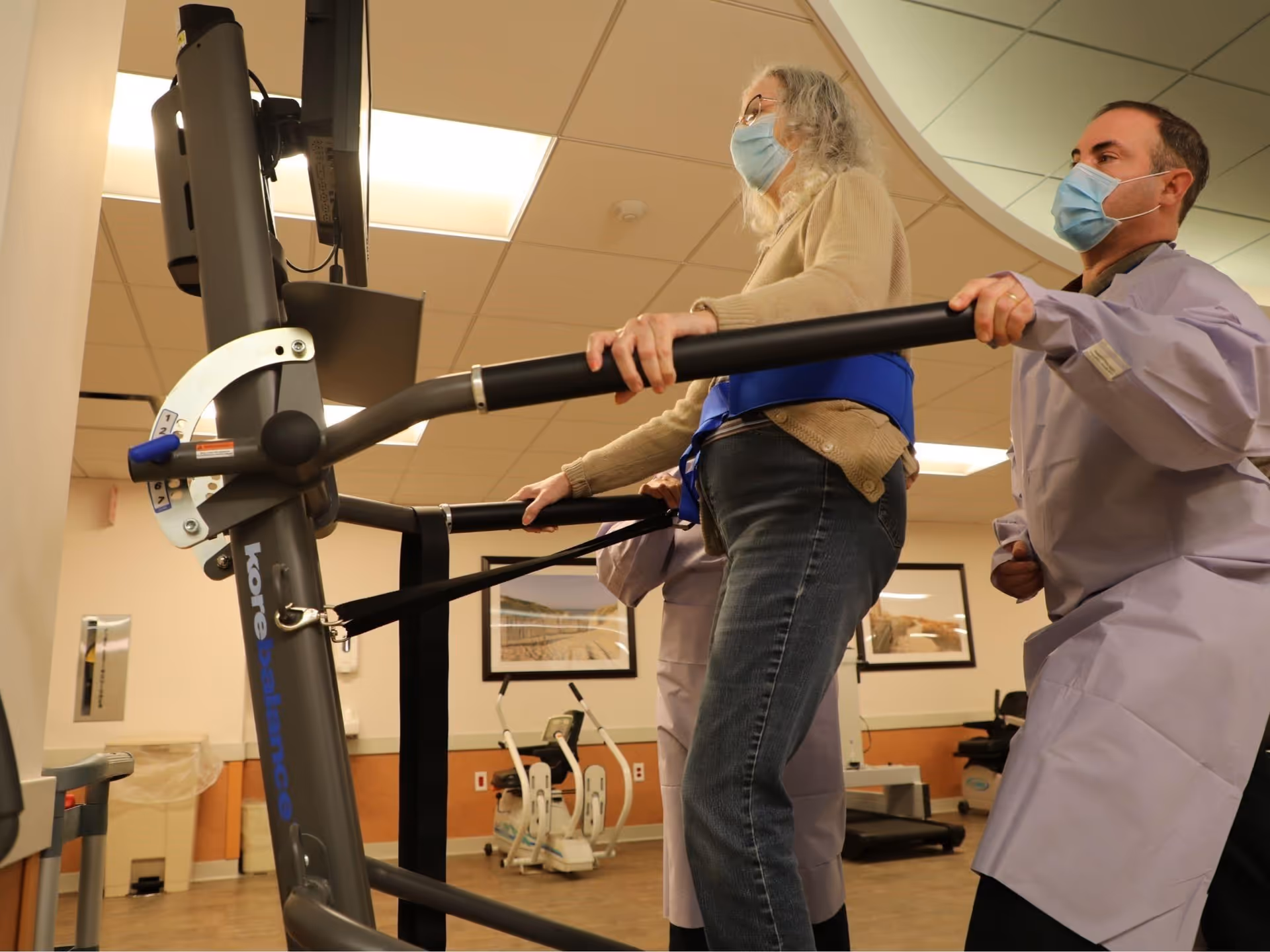 An elderly woman wearing a face mask is using a treadmill machine for rehabilitation while being assisted by a healthcare professional also wearing a face mask in a therapy room.