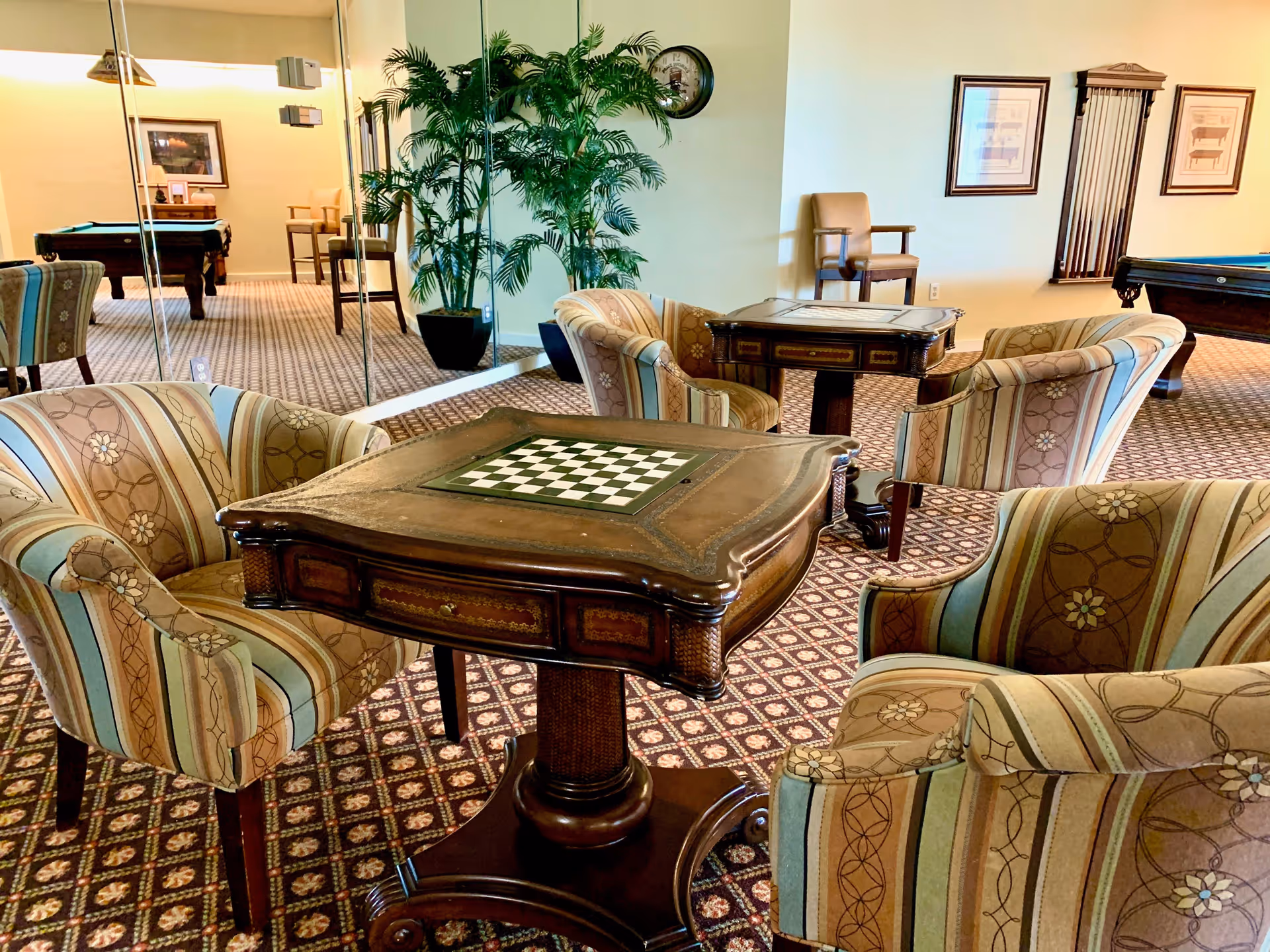 A cozy indoor lounge area with patterned carpet, two wooden game tables with built-in chess boards, and four upholstered armchairs with striped and floral designs. In the background, there is a large potted plant, a wall clock, framed pictures, a chair, and a pool table. A mirrored wall reflects part of the room.