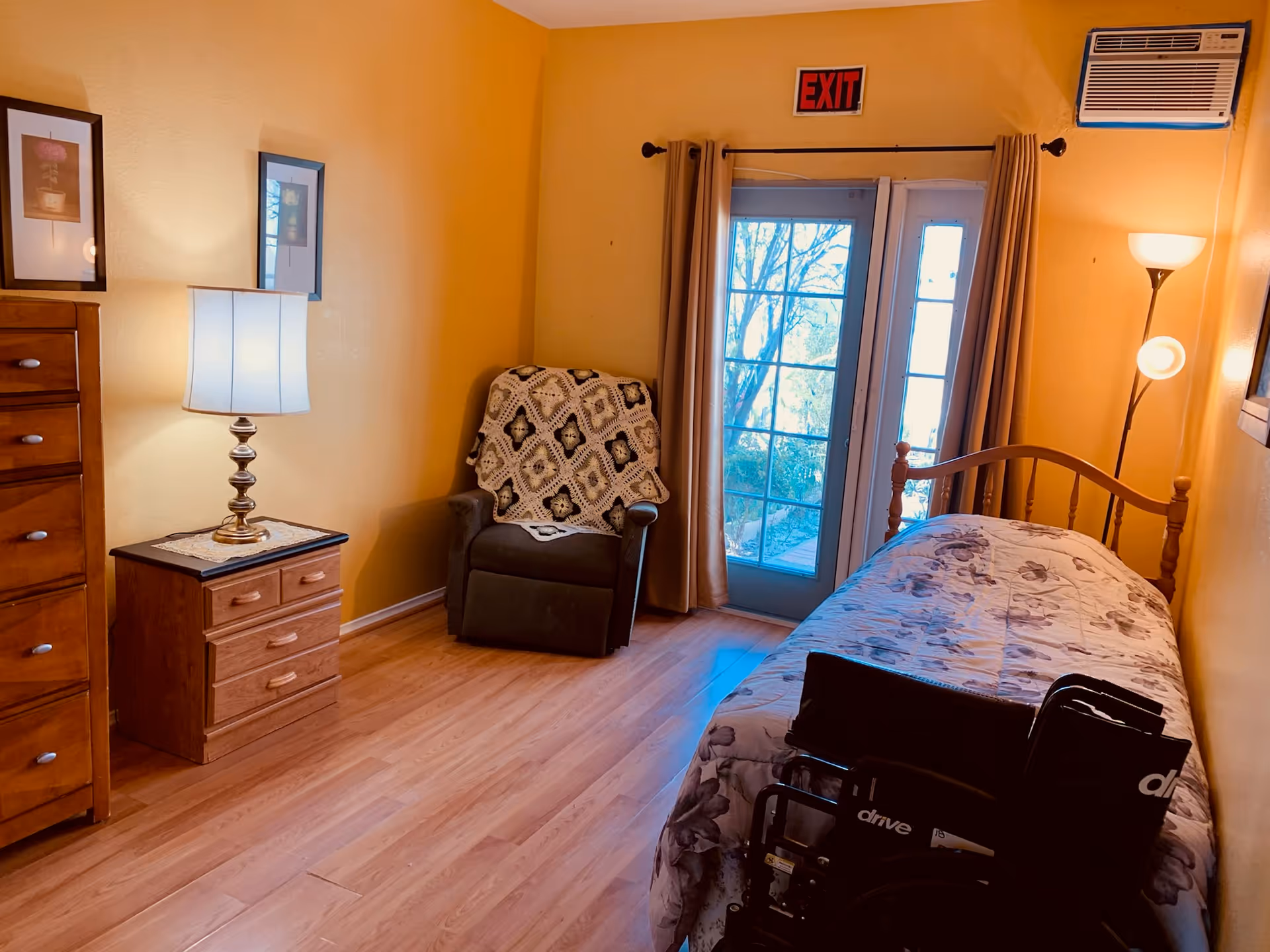 Yellow-walled bedroom with single bed, dresser, chair, wheelchair, and French doors.