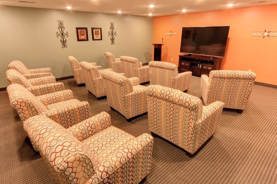 A cozy media room with two rows of patterned armchairs facing a large flat-screen TV mounted on an orange wall. The room has a carpeted floor, recessed ceiling lights, and decorative wall hangings on the adjacent green wall.