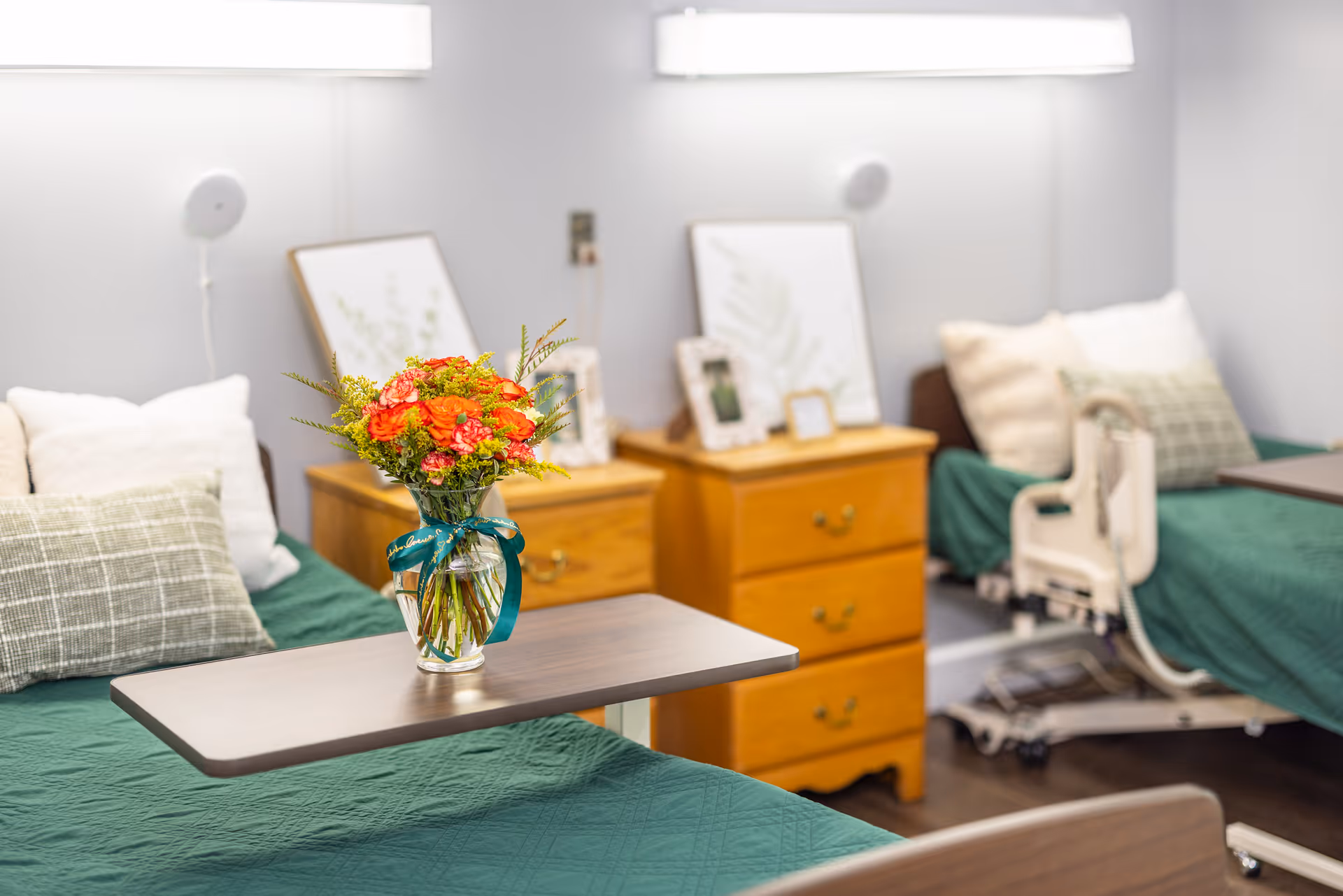 Shared patient bedroom with two hospital-style beds, green bedspreads, wooden nightstands, and a vase of flowers on an overbed table.