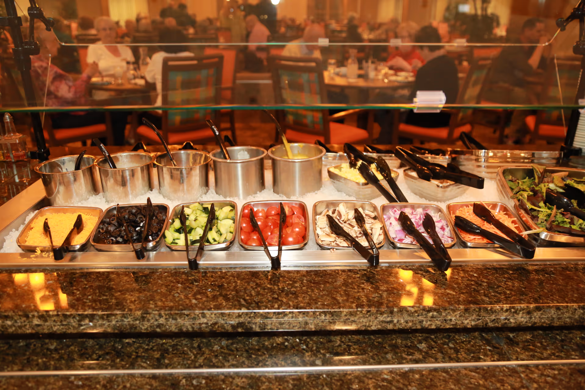 A salad bar with various fresh ingredients including shredded cheese, black olives, cucumber slices, cherry tomatoes, sliced mushrooms, chopped onions, shredded carrots, and leafy greens. Several metal containers hold dressings and other toppings, with tongs and serving spoons placed for self-service. In the background, people are seated at tables in a dining area.