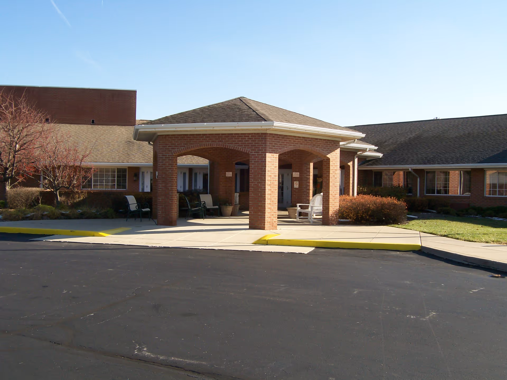 Exterior view of a brick building with a covered entrance supported by brick columns. There are chairs placed under the covered area and some bushes and trees around the building. The sky is clear and blue.