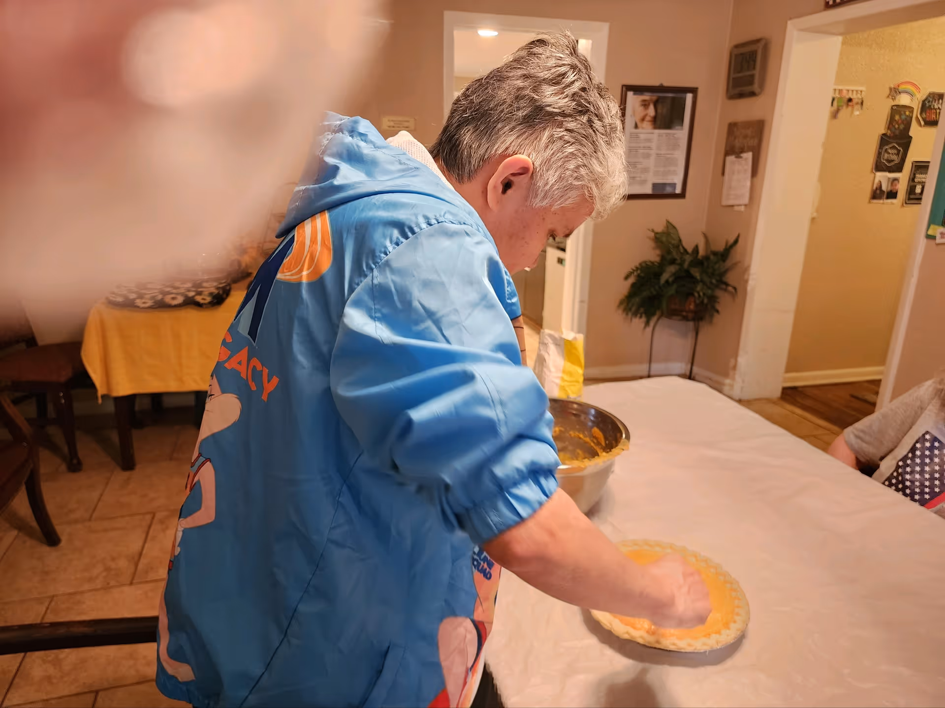 An elderly person wearing a blue jacket is preparing a pie on a table covered with a white tablecloth in a cozy indoor setting. There is a large metal mixing bowl with orange filling on the table. The room has beige walls, a plant in the corner, and various decorations on the walls.