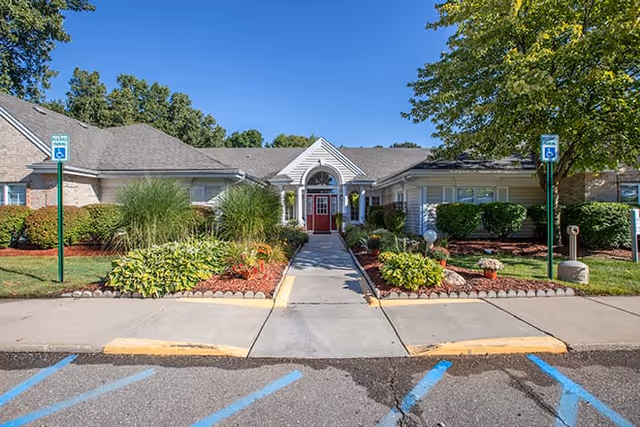 Front entrance of a single-story senior living building with a red door, landscaped walkway, and accessible parking spaces.