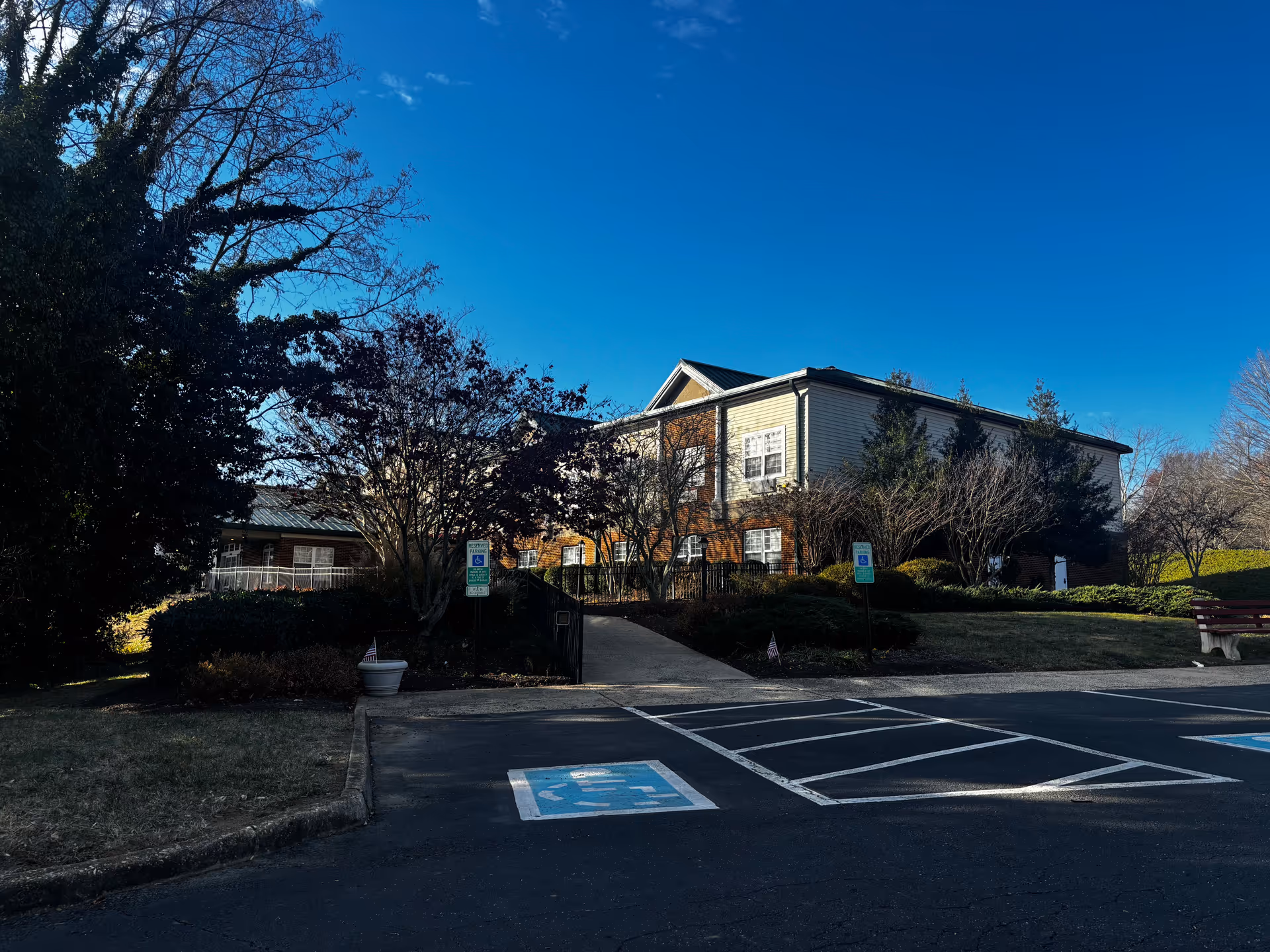 Exterior view of Commonwealth Senior Living at Stafford building with a clear blue sky, surrounded by trees and bushes. There is a paved parking area in the foreground with marked handicapped parking spaces and signs. A ramp leads up to the entrance of the building.