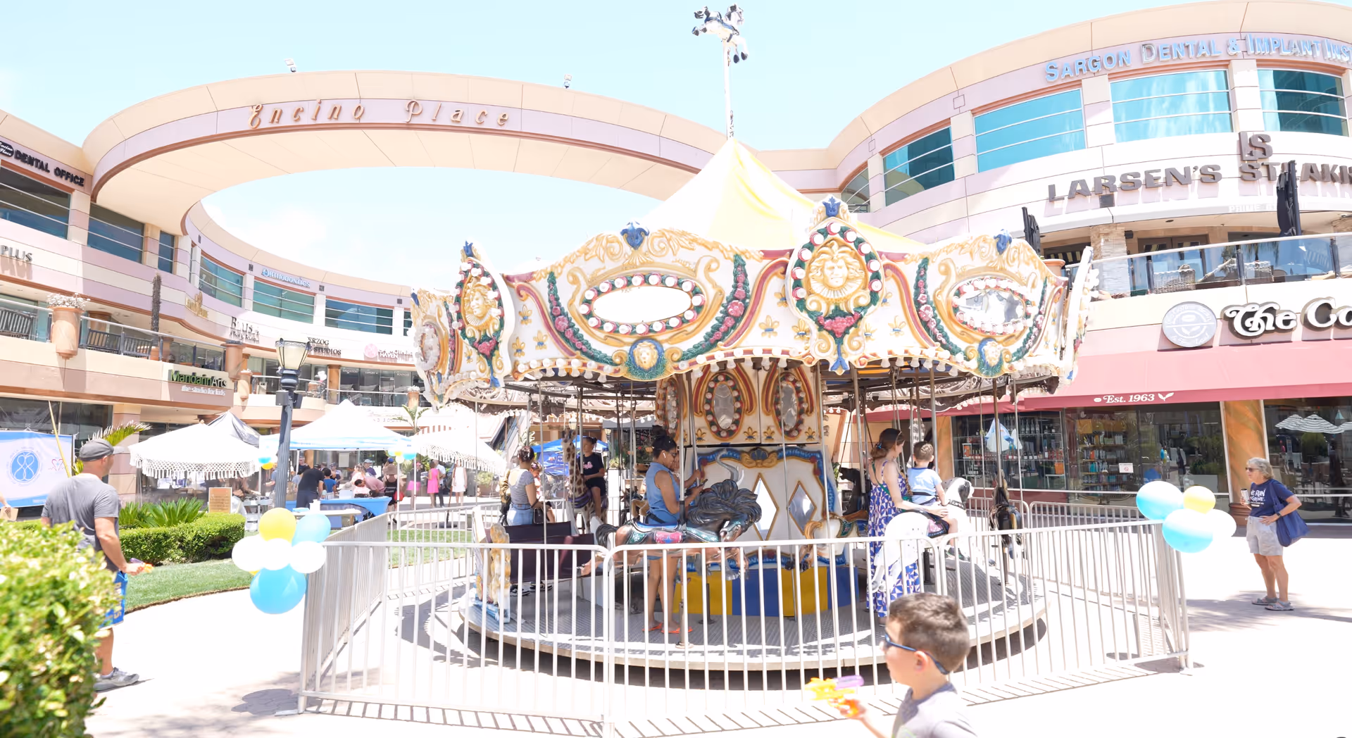 Colorful outdoor carousel in a sunny shopping plaza with storefronts and people nearby.