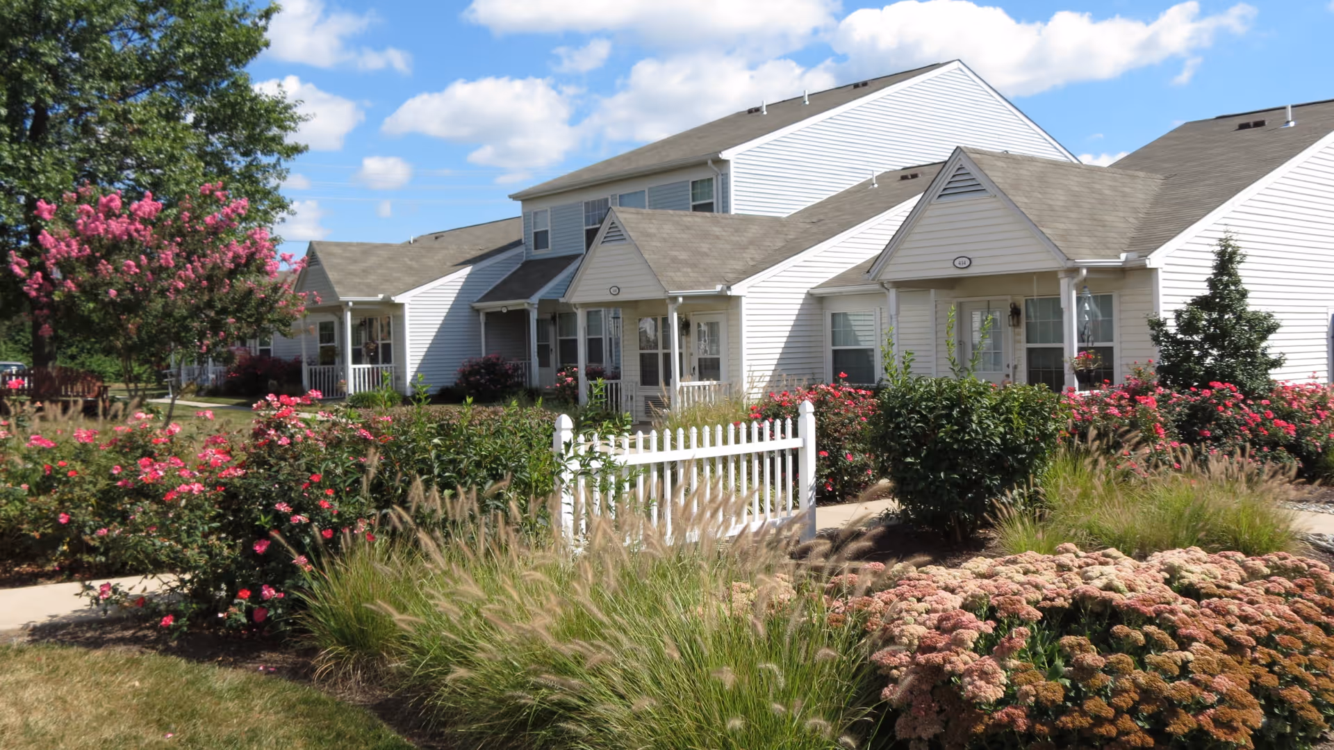 Front exterior of white-siding senior living cottages with a small picket fence, flowering shrubs, and manicured landscaping under a blue sky.
