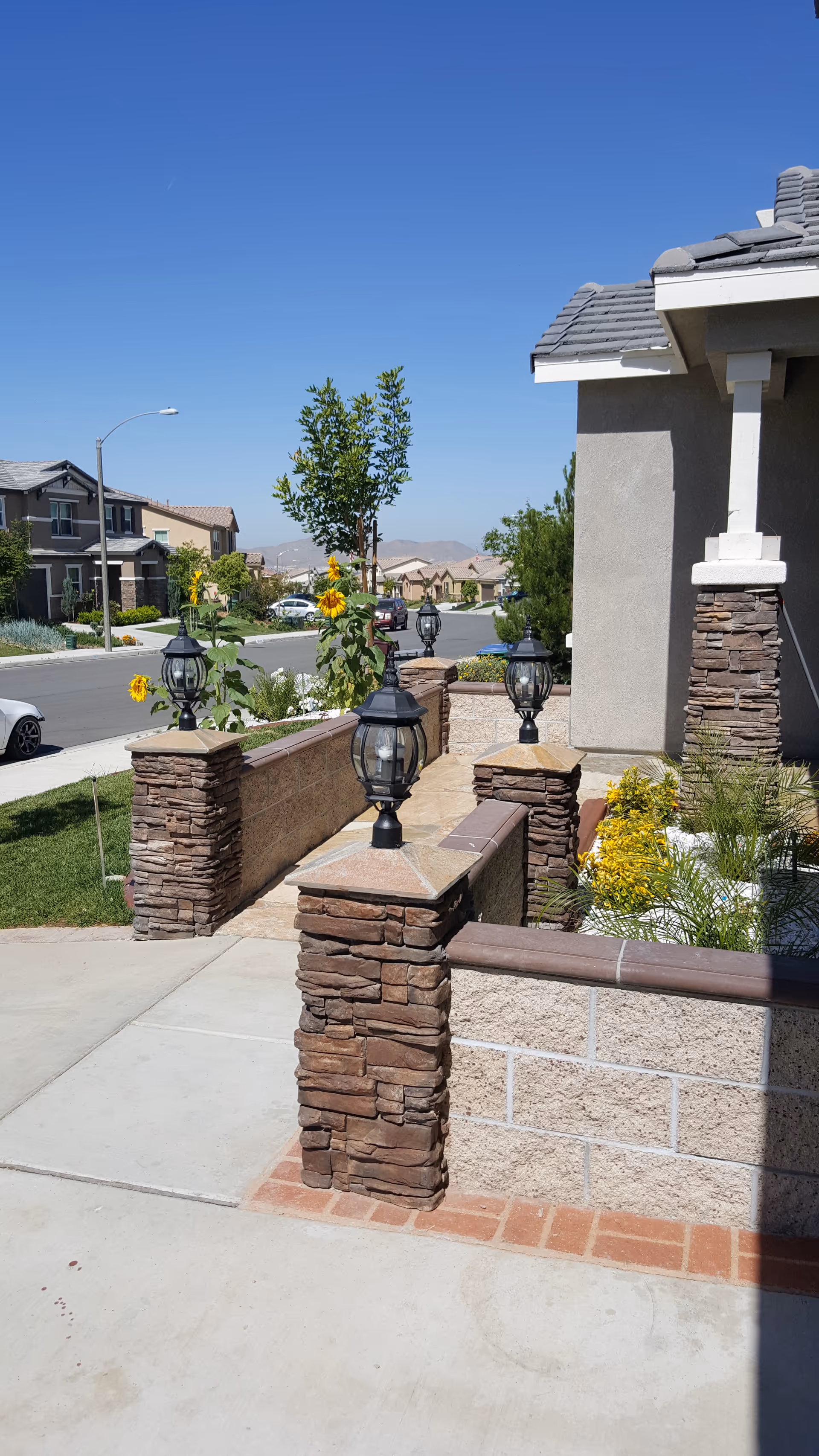 Stone-columned entrance with lanterns, walkway, sunflowers, and a sunny residential street.