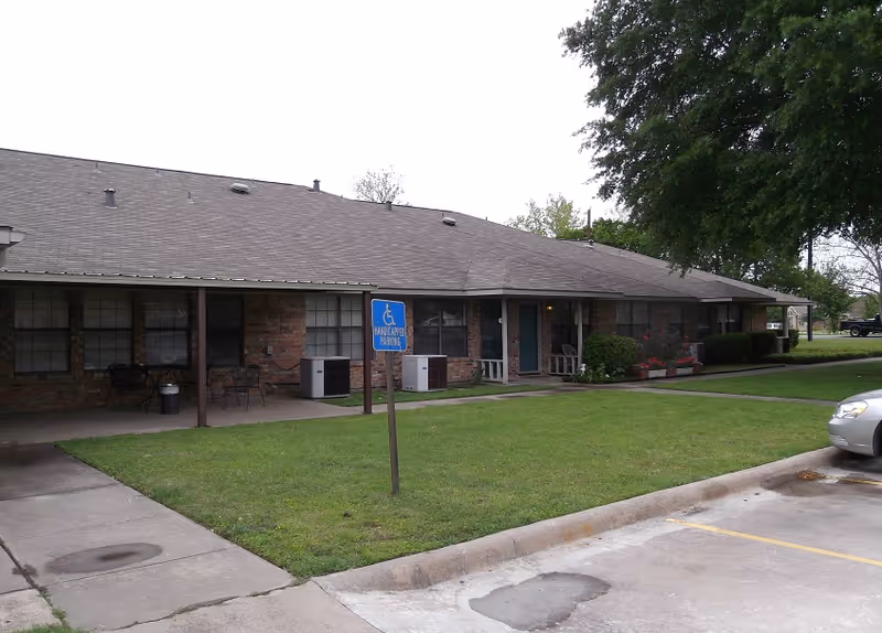 Exterior view of a single-story brick building with a covered porch area, several windows, and two blue doors. There is a grassy area in front with a handicapped parking sign, and a silver car is partially visible parked on the right side. Trees and shrubs surround the building.