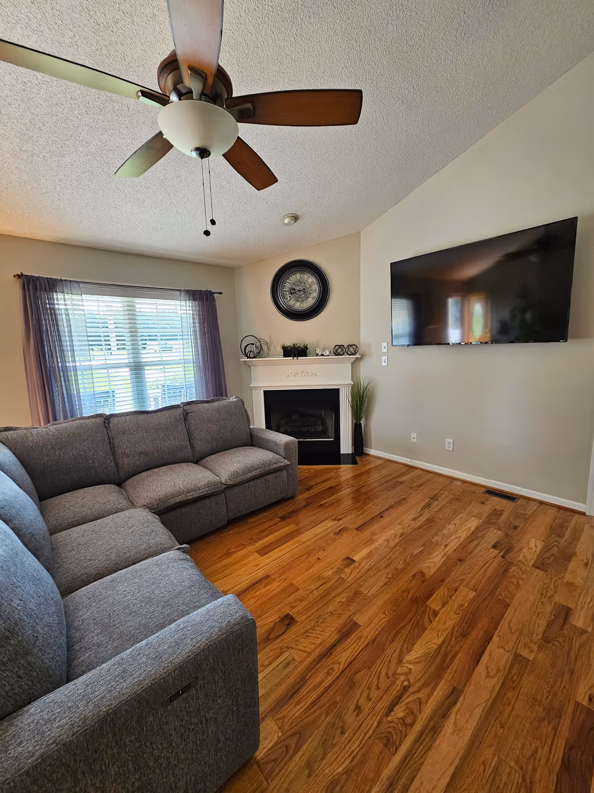 Living room with gray sectional sofa, fireplace mantle, TV, ceiling fan, and hardwood floors.