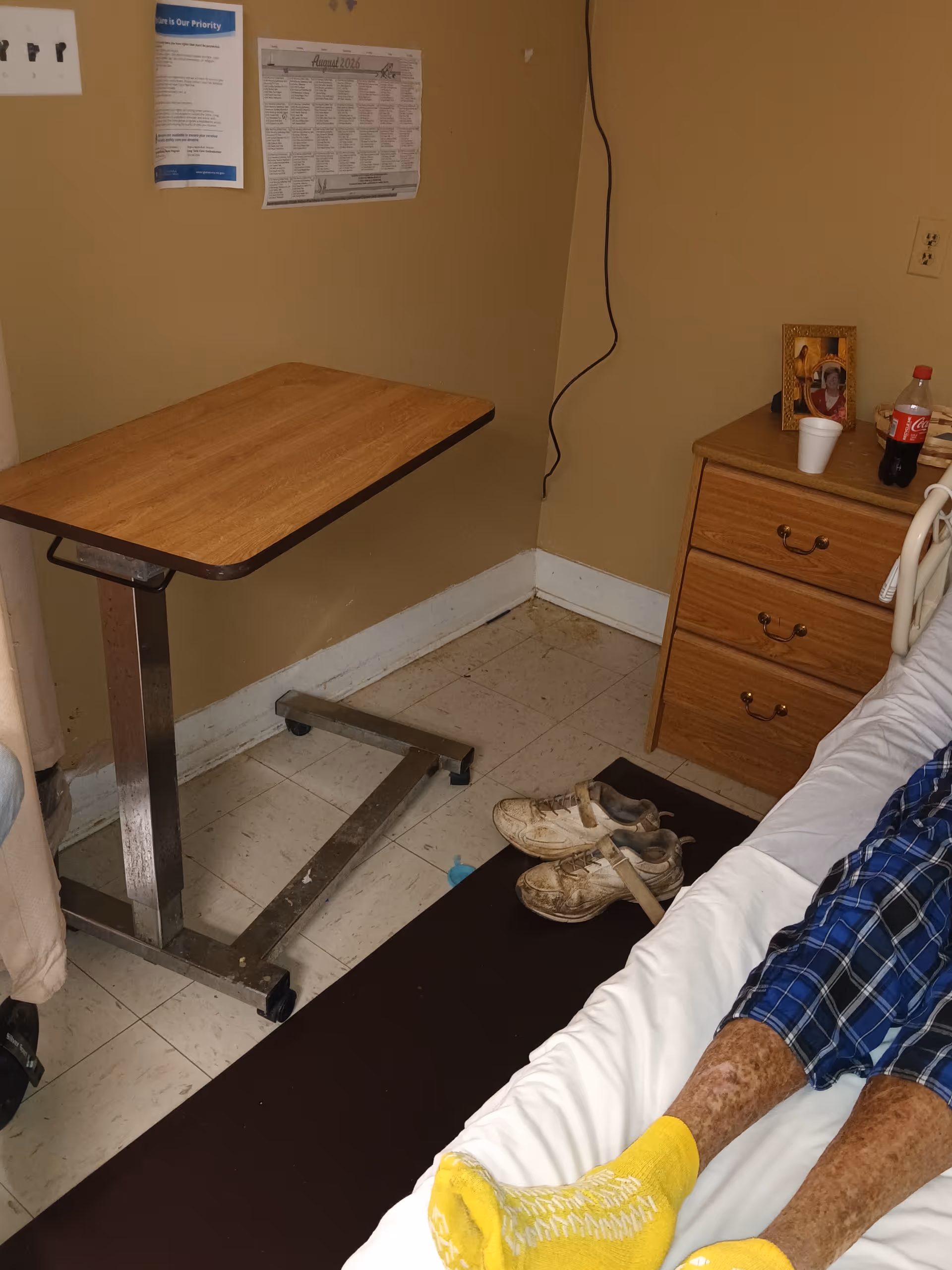 A small hospital-style room corner with a folding table, dresser, shoes, and a patient in bed.
