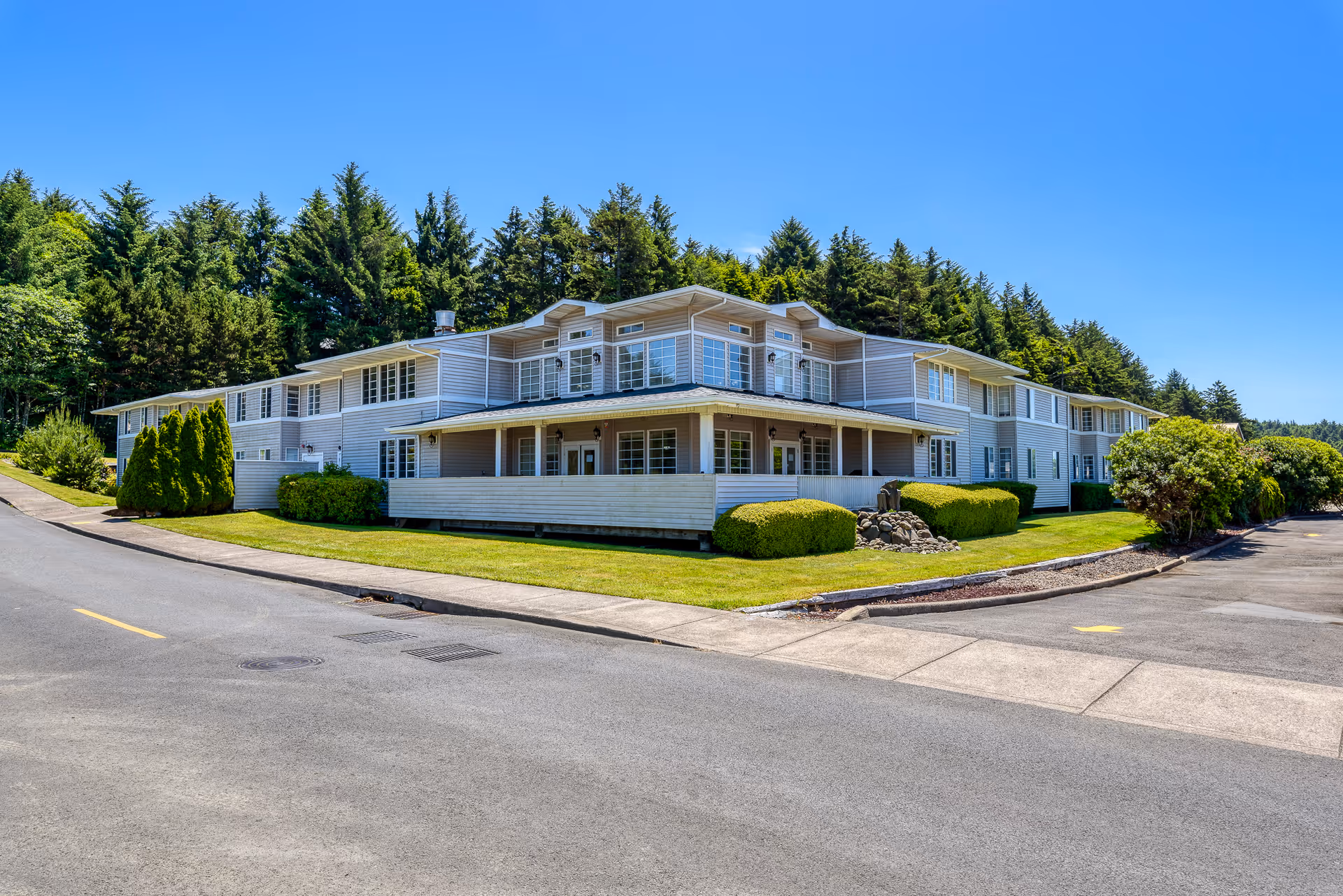 Exterior view of a two-story assisted living facility building with white siding, surrounded by well-maintained green lawns, bushes, and trees under a clear blue sky.