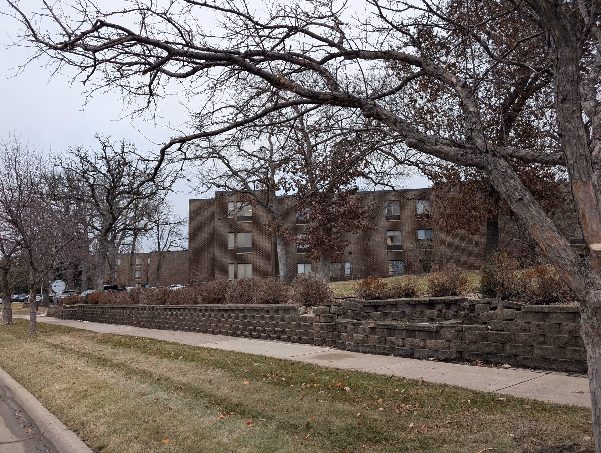 Brick three-story building set behind a tiered stone retaining wall with leafless trees and a sidewalk in front.