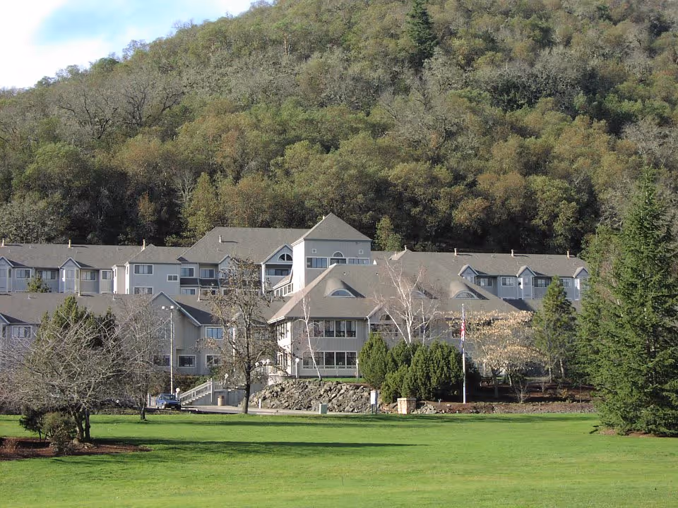 A large multi-story residential building surrounded by trees and greenery with a forested hill in the background under a partly cloudy sky.
