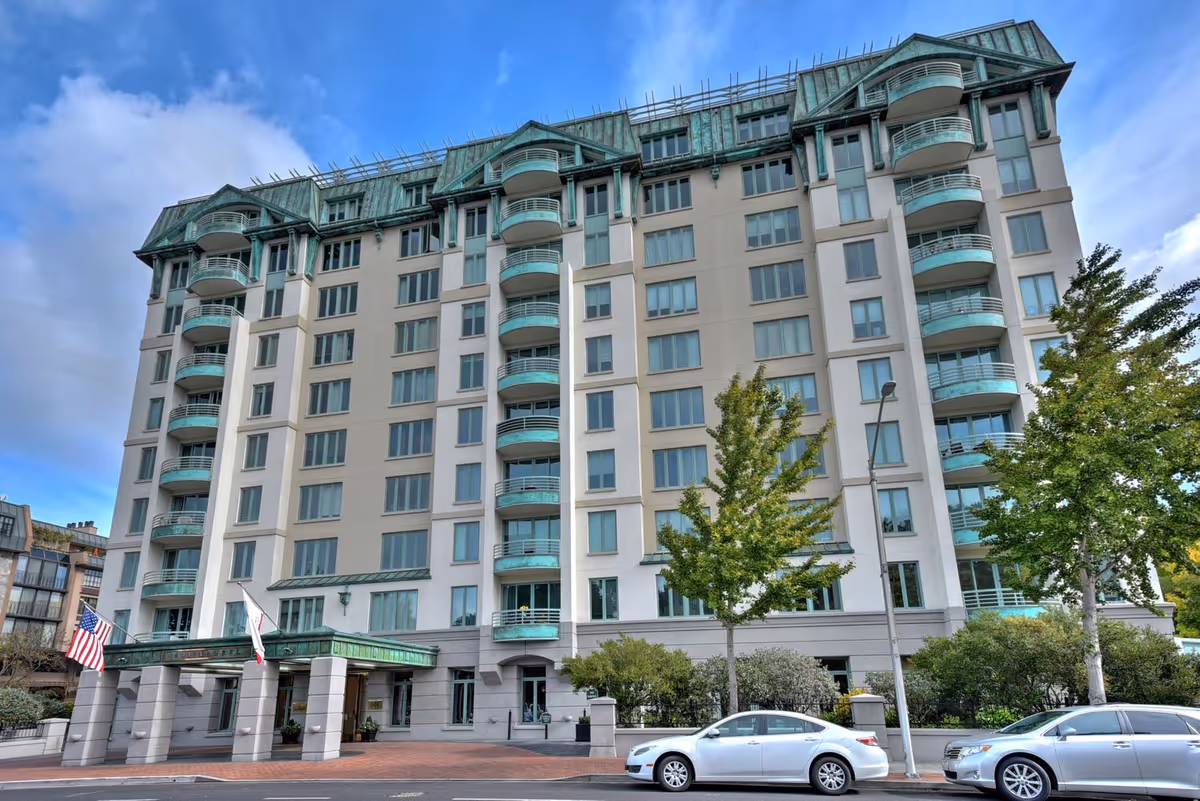 Tall modern senior living building with balconies, trees, and a covered entrance on a street.