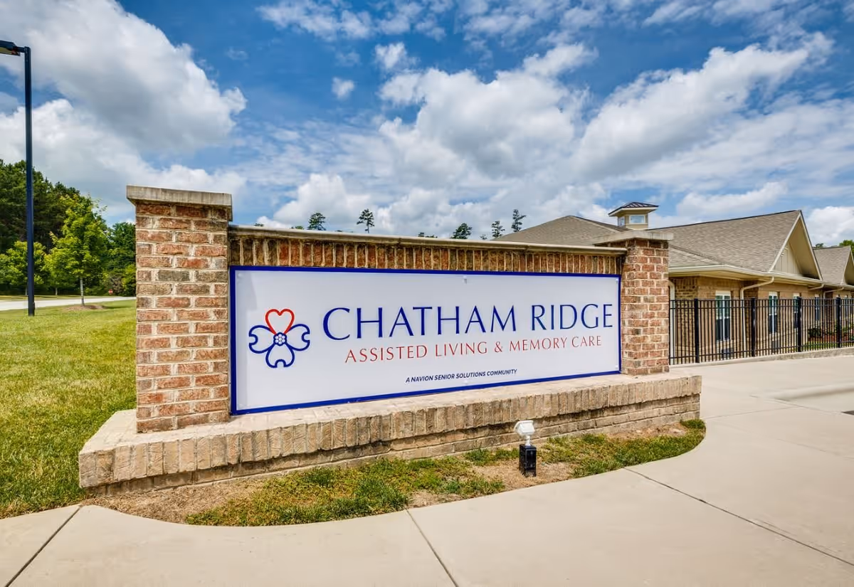Outdoor view of a brick sign for Chatham Ridge Assisted Living & Memory Care with a building and a black metal fence in the background under a partly cloudy blue sky.