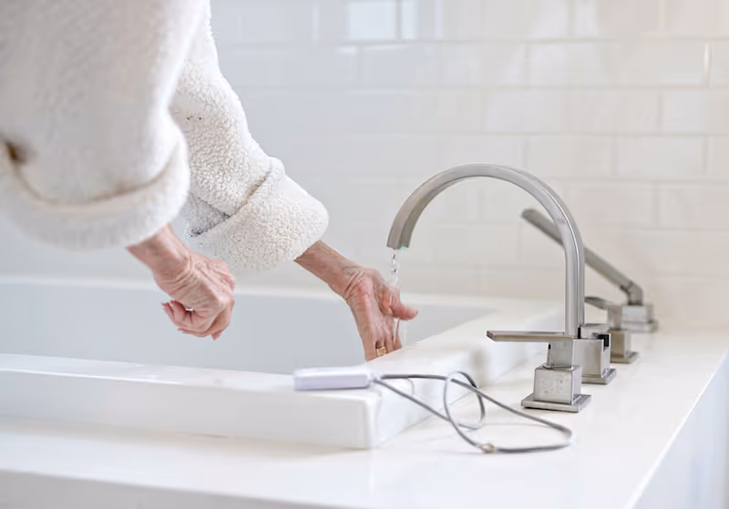 Elderly person washes hands at a white bathroom sink with chrome faucet.