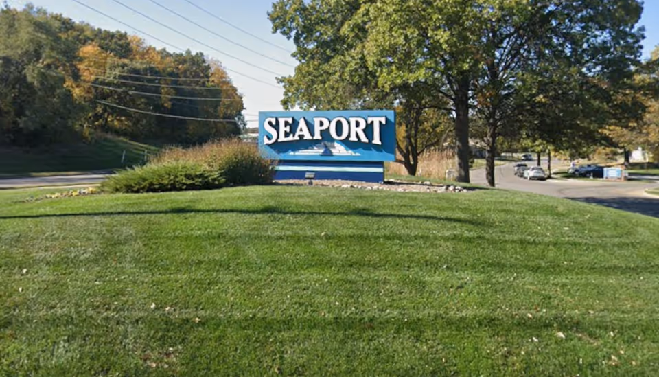 Seaport sign on a grassy mound with trees and a road behind.