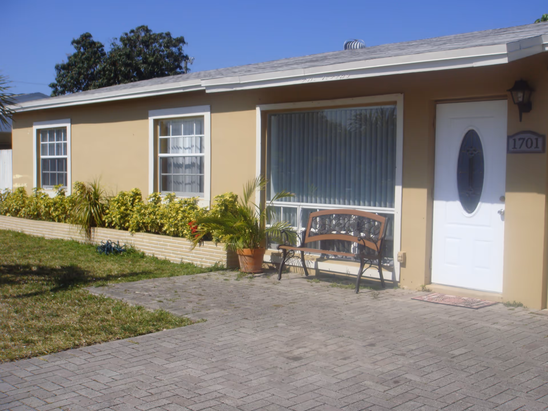Single-story beige building front with a white door numbered 1701, large window, a bench and potted plants beside a brick-paved driveway.