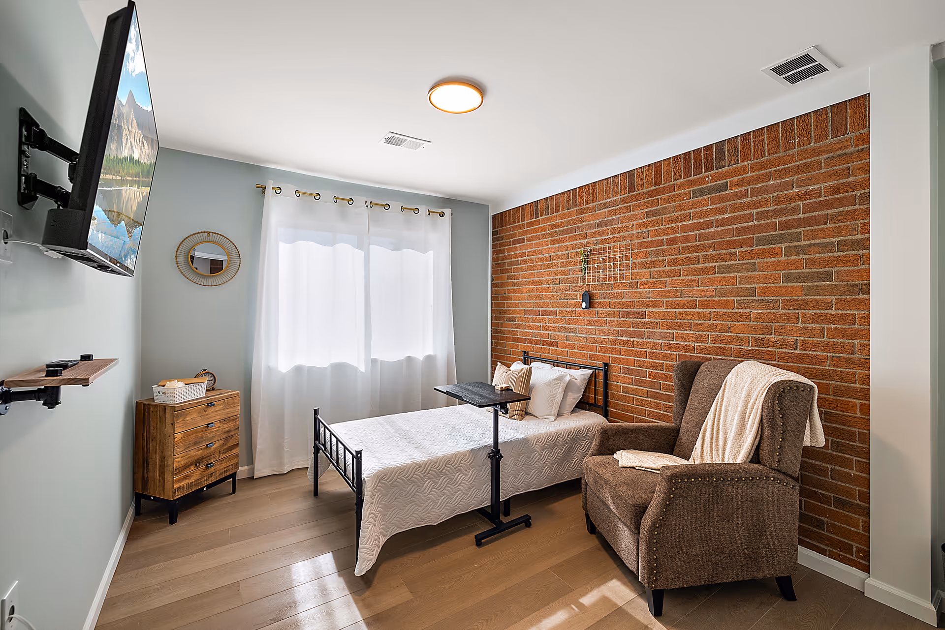 Bright bedroom with brick accent wall, bed, chair, dresser, and wall-mounted TV.