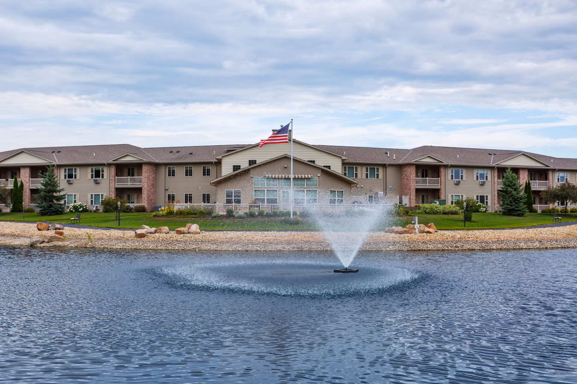 View of a senior living facility building with beige siding and brick accents, situated behind a pond with a water fountain spraying water upwards. An American flag is flying on a flagpole in front of the building, and there is a grassy area with trees and benches around the pond under a partly cloudy sky.