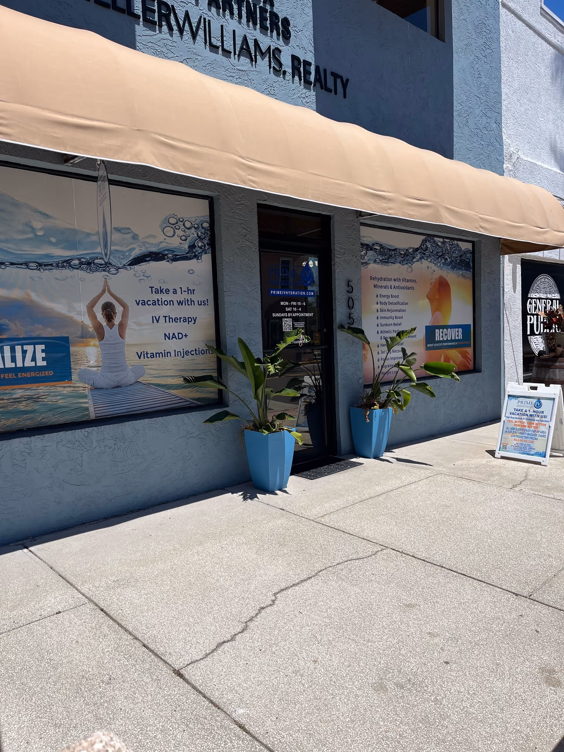 Front of Prime IV Hydration & Wellness storefront with beige awning and blue planters.