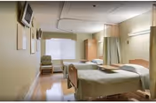 A sunlit shared patient room with two beds, privacy curtains, and light furniture.