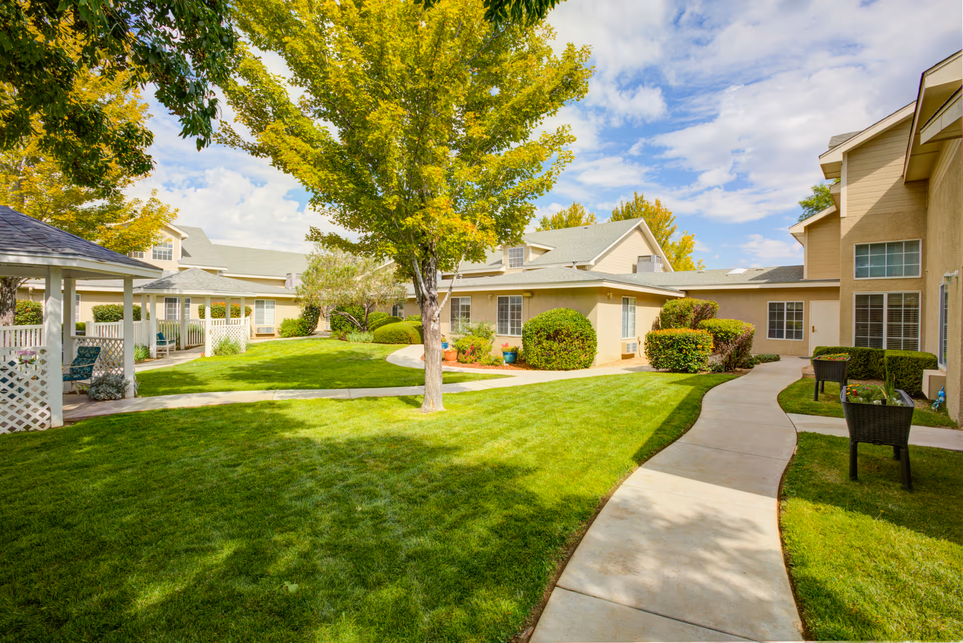 A sunny outdoor courtyard at Avista Senior Living Albuquerque featuring a well-maintained green lawn, a paved walkway, several trees, bushes, and beige buildings with multiple windows surrounding the area. There are also a few black chairs and a small gazebo visible.