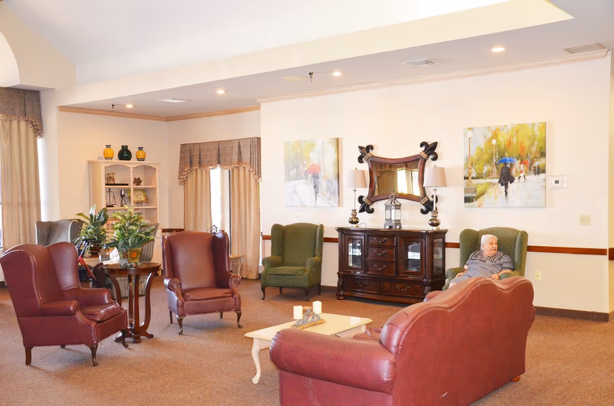 Senior living lounge with red leather chairs, green armchairs, and a resident seated nearby.