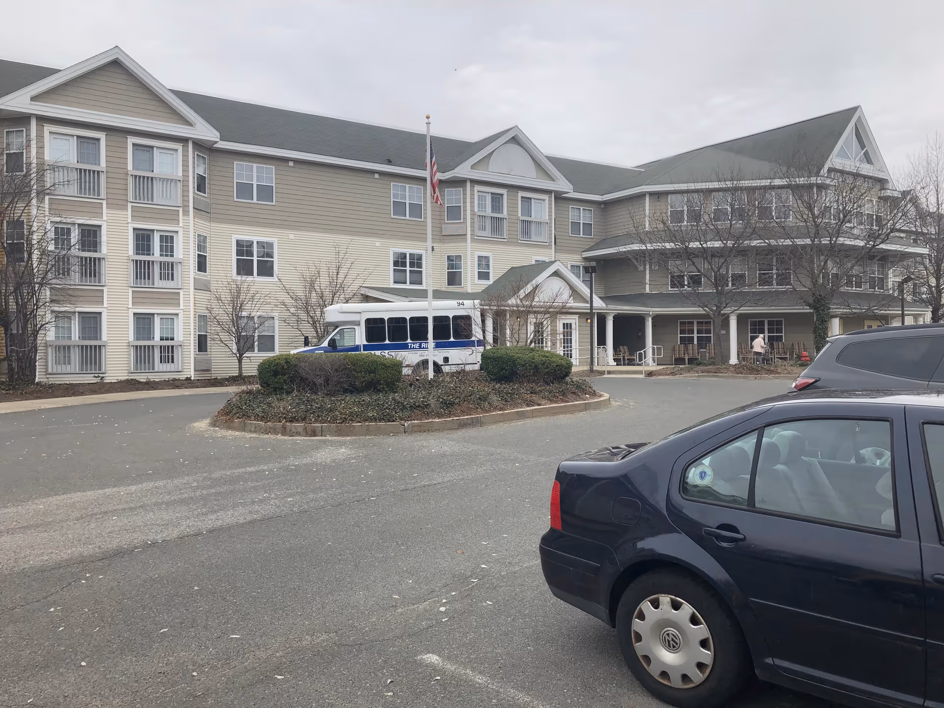Exterior view of a multi-story senior living facility with a bus and parked cars.