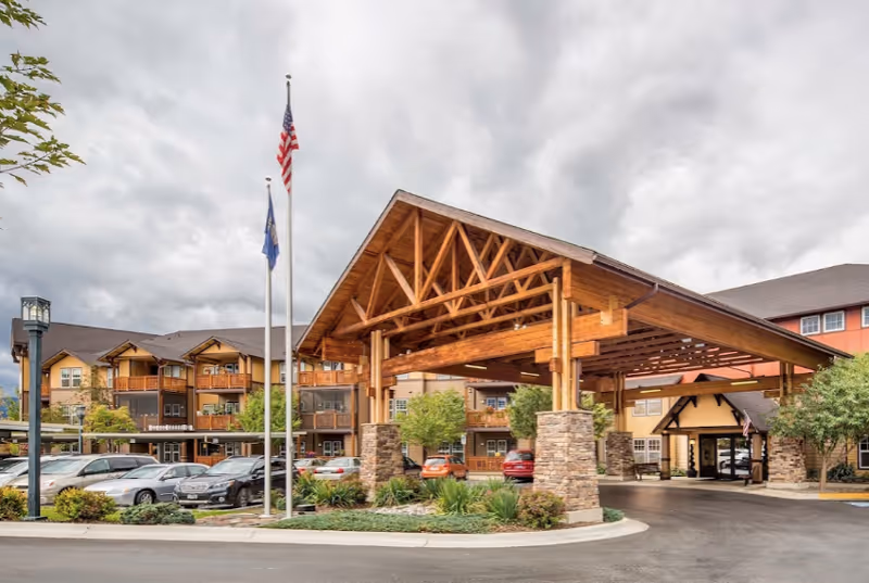 Exterior view of The Springs at Missoula senior living facility showing a large wooden porte-cochere entrance supported by stone pillars, with multiple parked cars and two flagpoles displaying the American flag and another flag. The building has multiple stories with balconies and a mix of stone and wood siding under a cloudy sky.