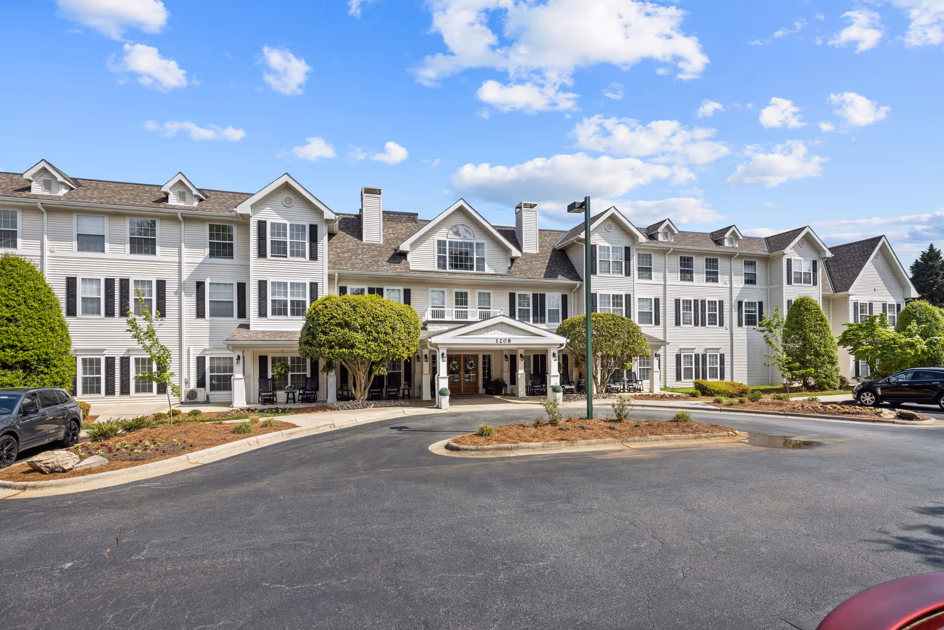 Front entrance of a three-story white residential building with a covered porte-cochere, circular driveway, parked cars, and landscaped grounds under a blue sky.