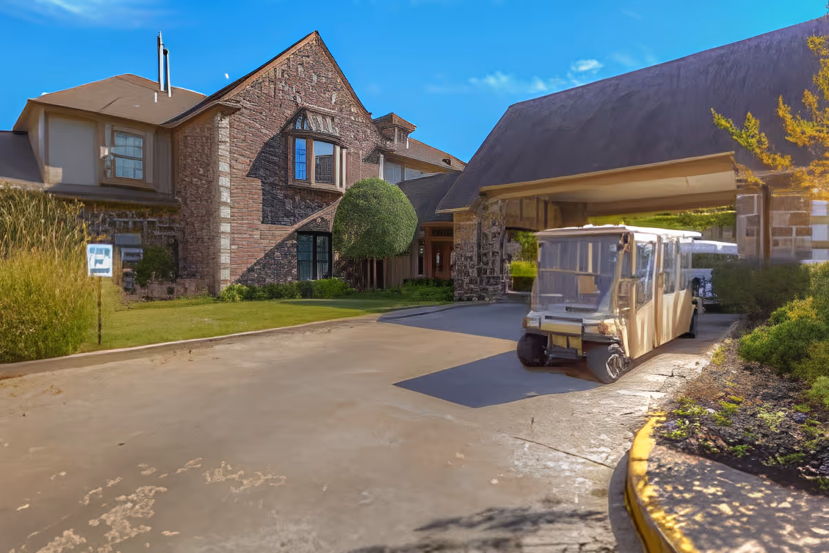 Exterior view of a large brick and stone building with a covered driveway entrance. A golf cart or small shuttle vehicle is parked under the covered area. The sky is clear and blue, and there is greenery including bushes and a tree around the building.