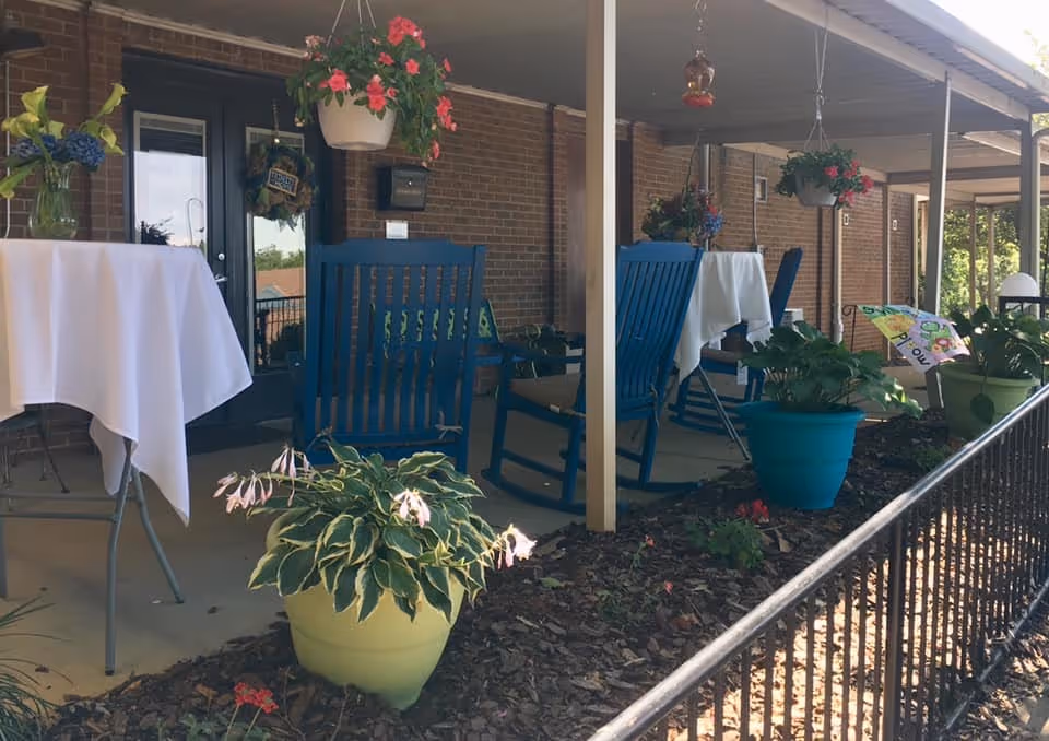 Brick exterior porch with blue rocking chairs, tables, and hanging plants on a shaded outdoor patio.