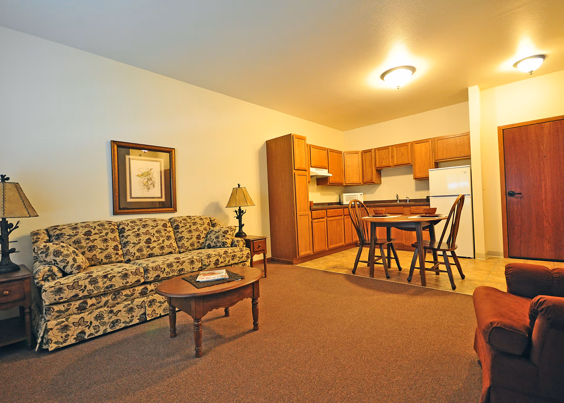Interior view of a living area in an assisted living facility featuring a floral patterned sofa with side tables and lamps, a wooden coffee table, a small dining table with chairs, and a kitchen area with wooden cabinets, a refrigerator, and a stove. The room has warm lighting and a closed wooden door.