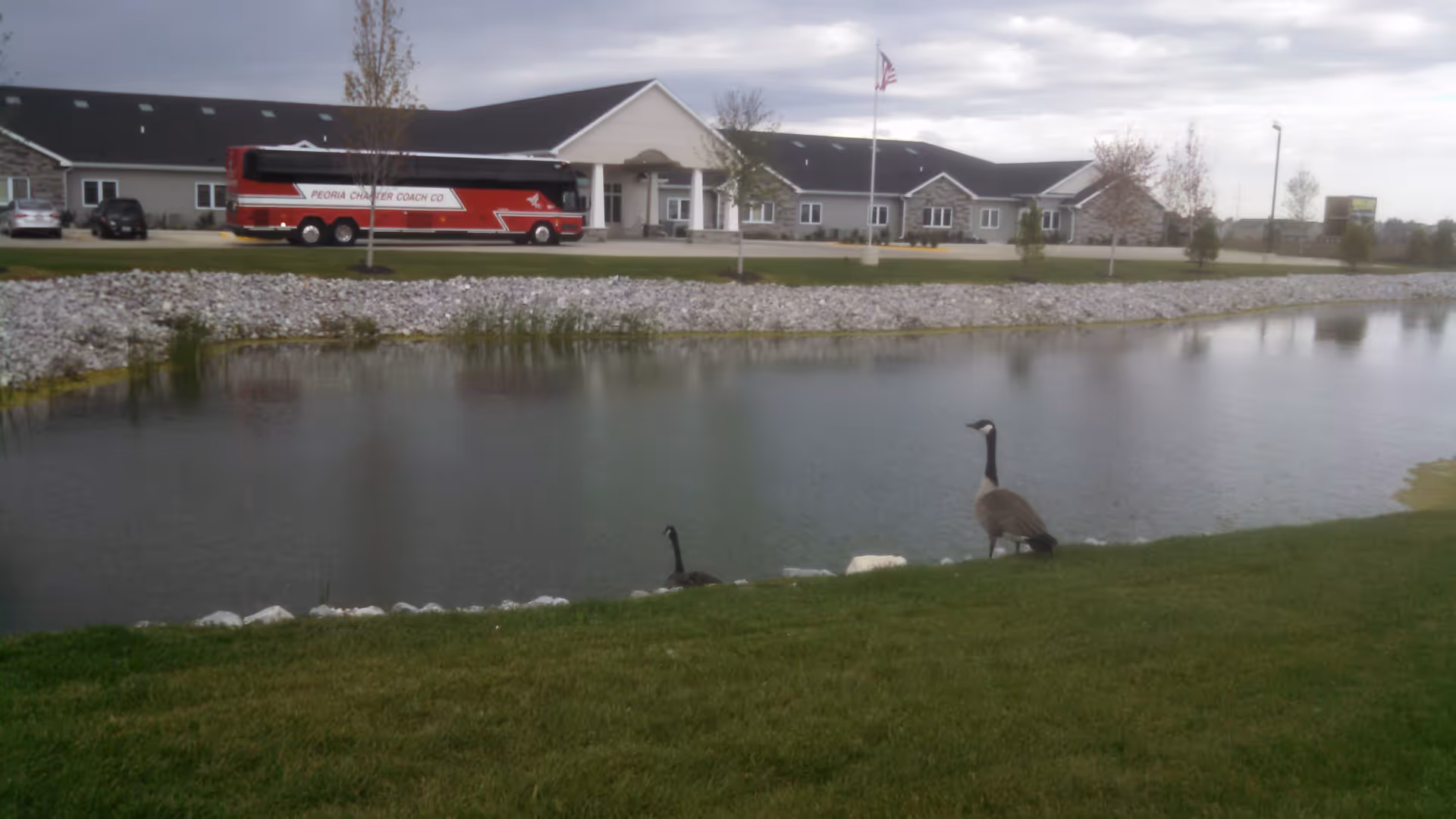View of Villas of Holly Brook Assisted Living facility in Washington, IL, showing a large building with a peaked roof, an American flag on a flagpole, a red and white Peoria Charter Coach bus parked in front, a pond with two geese near the grassy edge, and a cloudy sky.