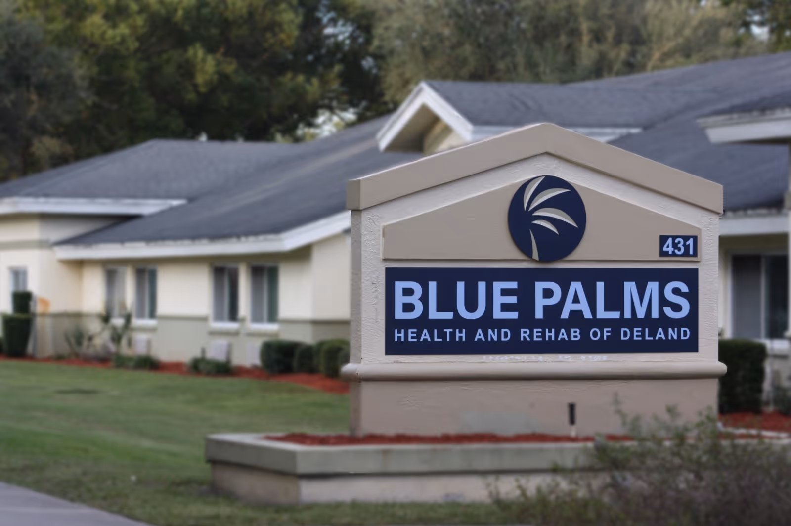 Outdoor view of the Blue Palms Health and Rehab of Deland facility sign with a beige building and green lawn in the background.
