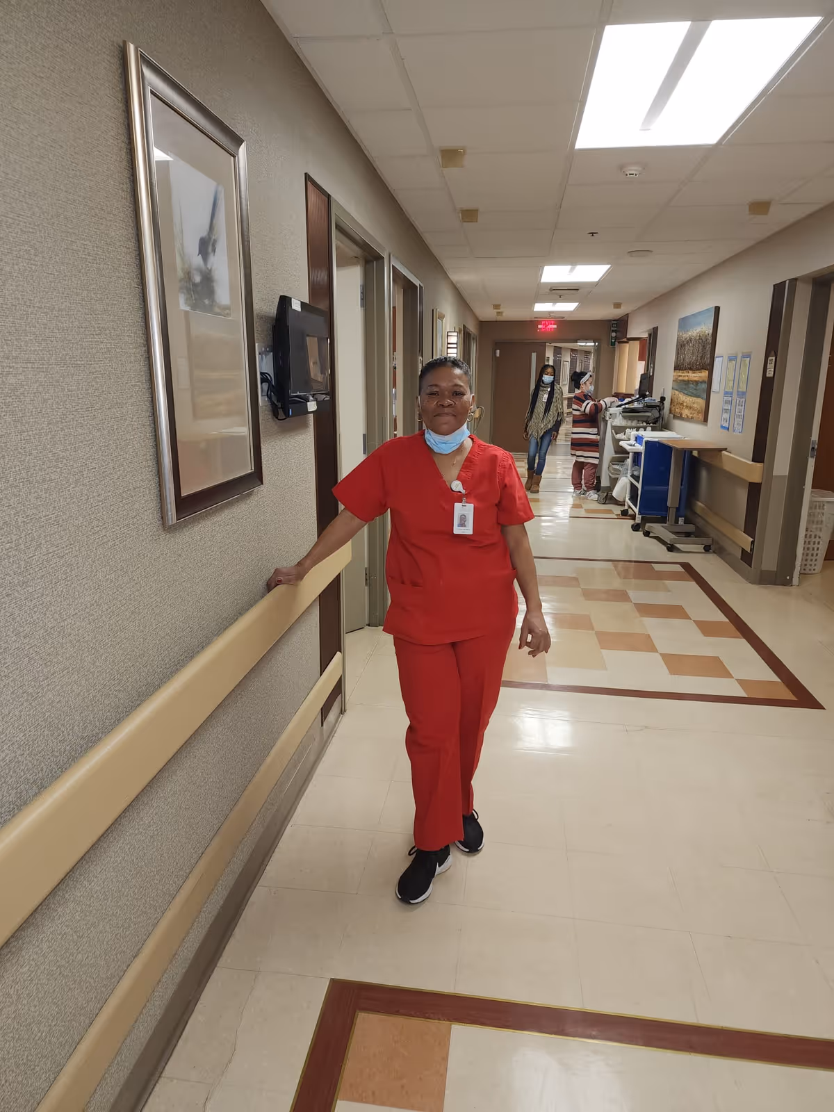 A healthcare worker in red scrubs stands in a well-lit hallway of a healthcare facility, leaning against a handrail. The worker is wearing a face mask pulled down below the chin and an ID badge. In the background, two other people, one wearing a mask, are visible further down the hallway near medical equipment and doors. The walls are decorated with framed artwork.