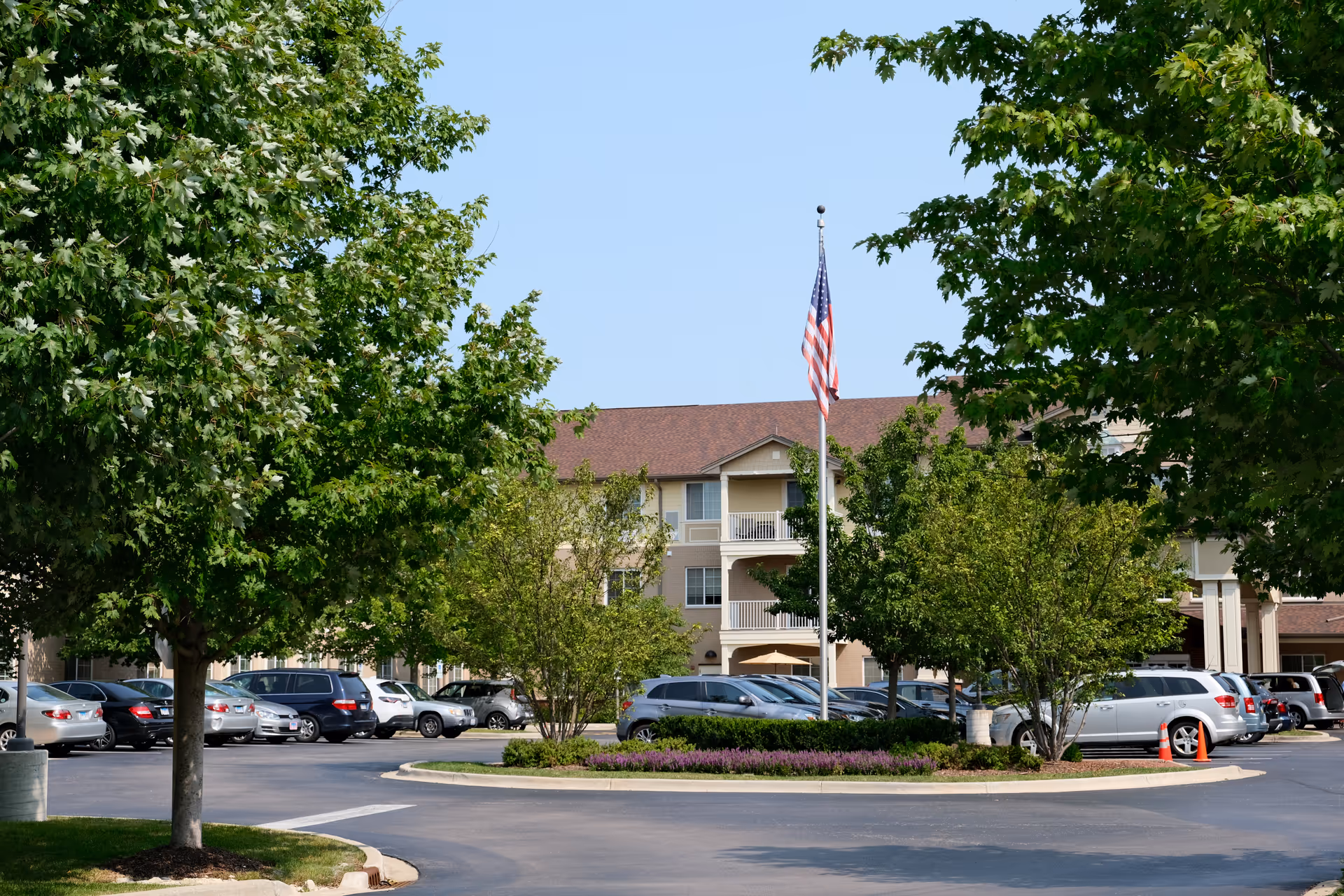 Front exterior of a three-story senior living building framed by trees with a parking lot and an American flag.