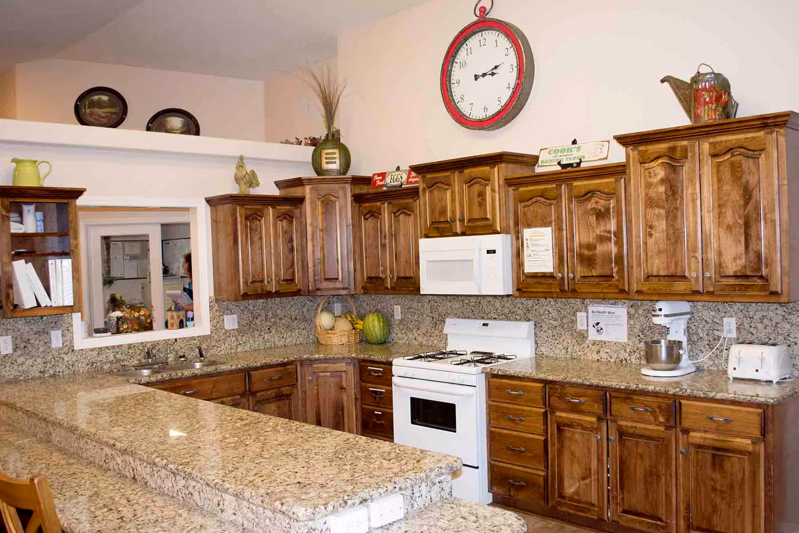 Bright communal kitchen with granite countertops, wooden cabinets, a white stove and microwave, and a large wall clock above.