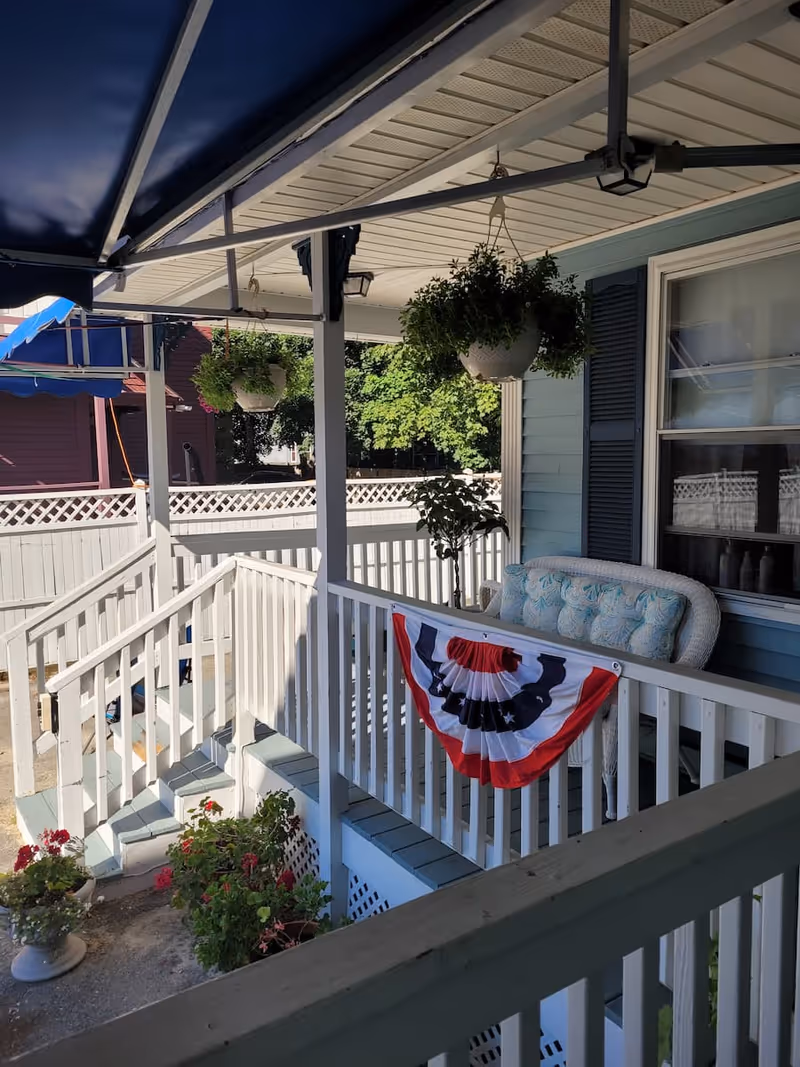 Front porch with white railing, hanging plants, a cushioned bench and a red-white-blue bunting.