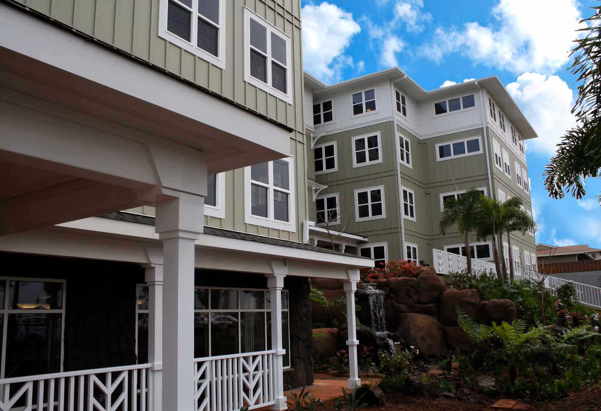 Exterior view of a multi-story light-green building with white trim, covered porch columns, and landscaped rocks with a small waterfall.