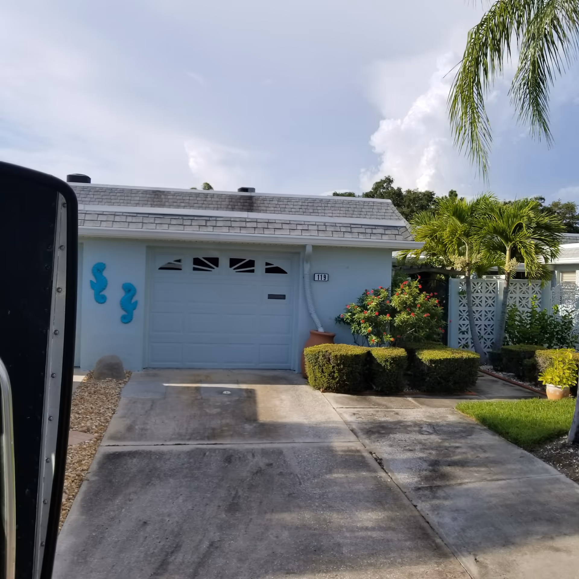 Front exterior of a light blue facility with a closed garage door, driveway, and tropical plants.