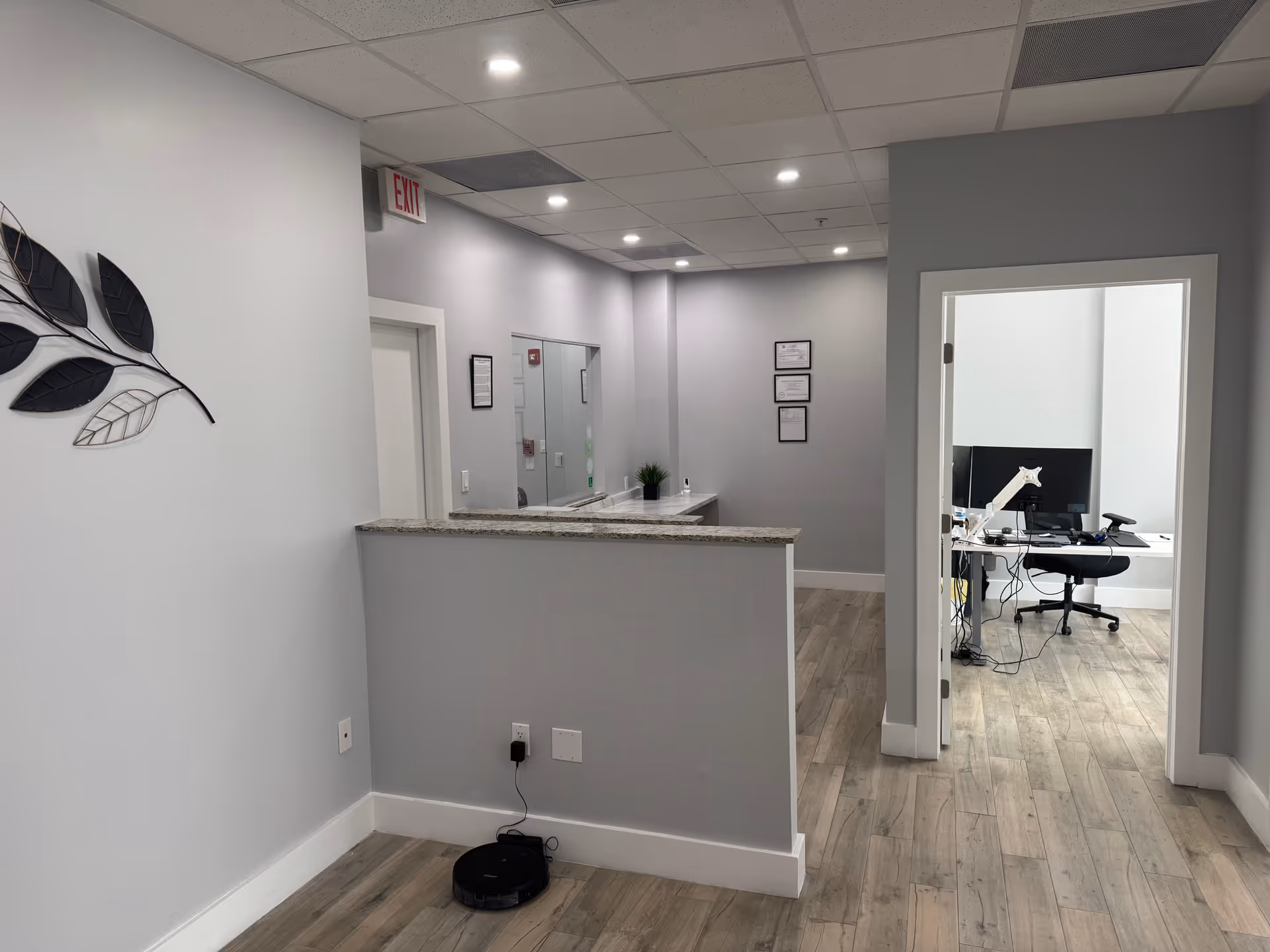Reception area with a counter, gray walls, exit sign, and an office doorway.
