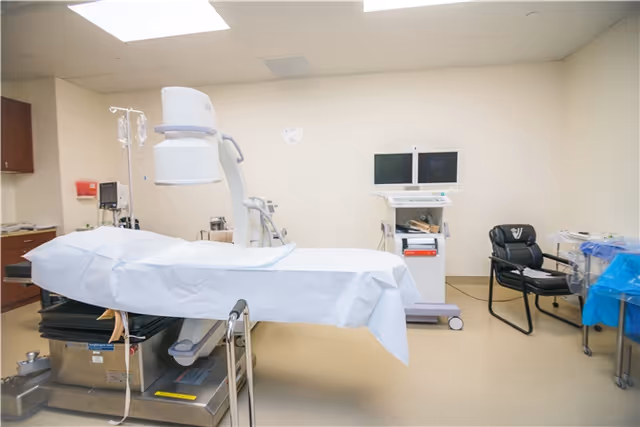 Medical procedure room with a covered operating table, overhead light, monitors, and equipment.