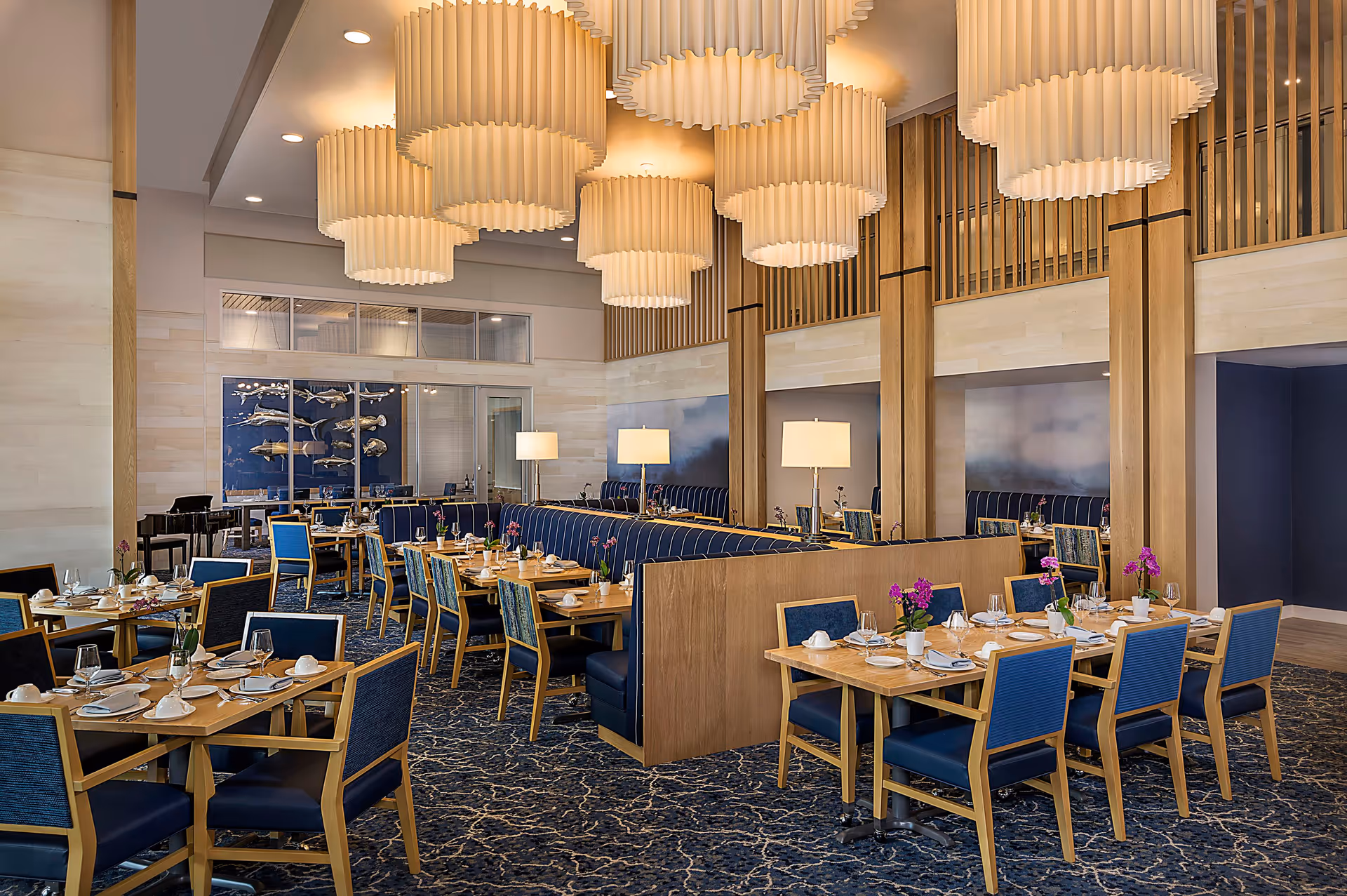 A spacious dining room with wooden tables and blue cushioned chairs, set with white plates, cups, and silverware. The room features large, modern pendant lights hanging from the ceiling, wooden paneling, and a patterned blue carpet. There are decorative purple flowers on the tables and lamps on a central divider. The walls have artistic decor and large windows.