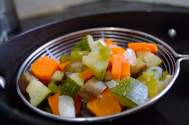 Colorful chopped vegetables in a metal colander over a pot in a kitchen.